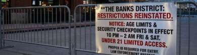 Metal barricades at The Banks district displaying a sign about reinstated age restrictions and security checkpoints.