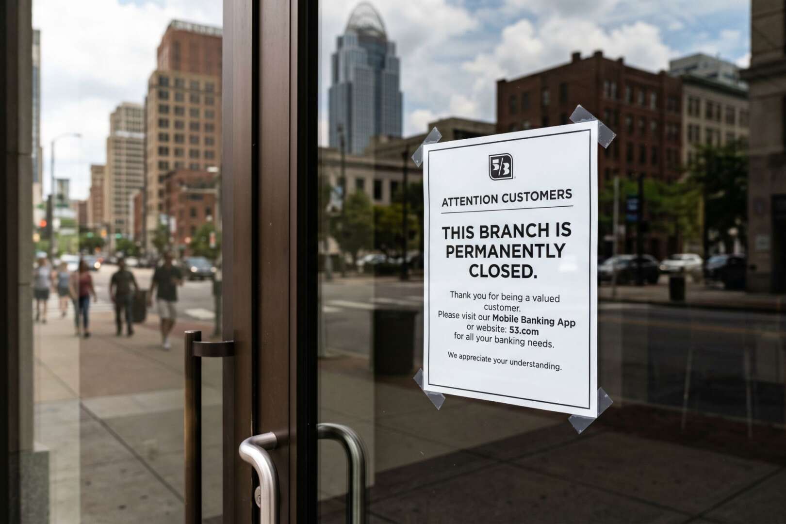 A sign on a glass door reading 'This branch is permanently closed' with the downtown Cincinnati skyline in the background, illustrating the local shift to digital banking.