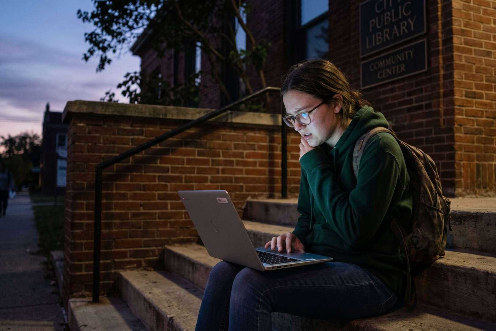 A student sits on the steps of a Cincinnati public library at dusk, using a laptop illuminated by the screen's glow to illustrate the digital divide and internet access gaps.