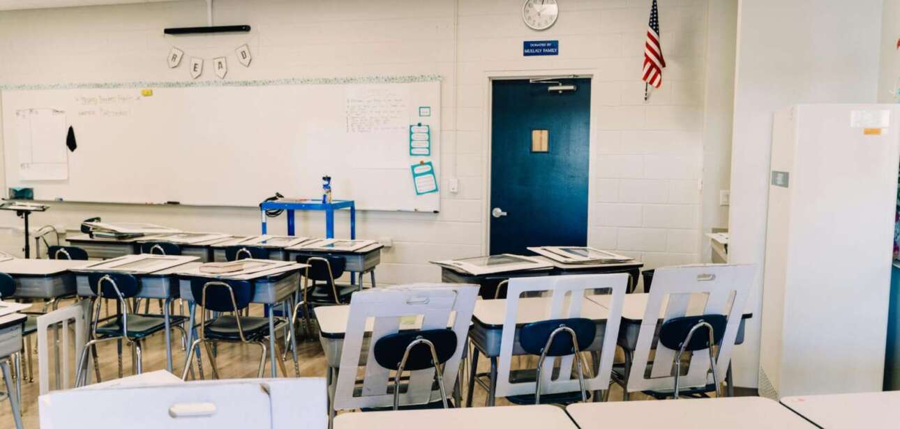 An empty, modern classroom at Cincinnati Public Schools with rows of white desks and chairs, a whiteboard, and an American flag, representing potential budget cuts or school closures.