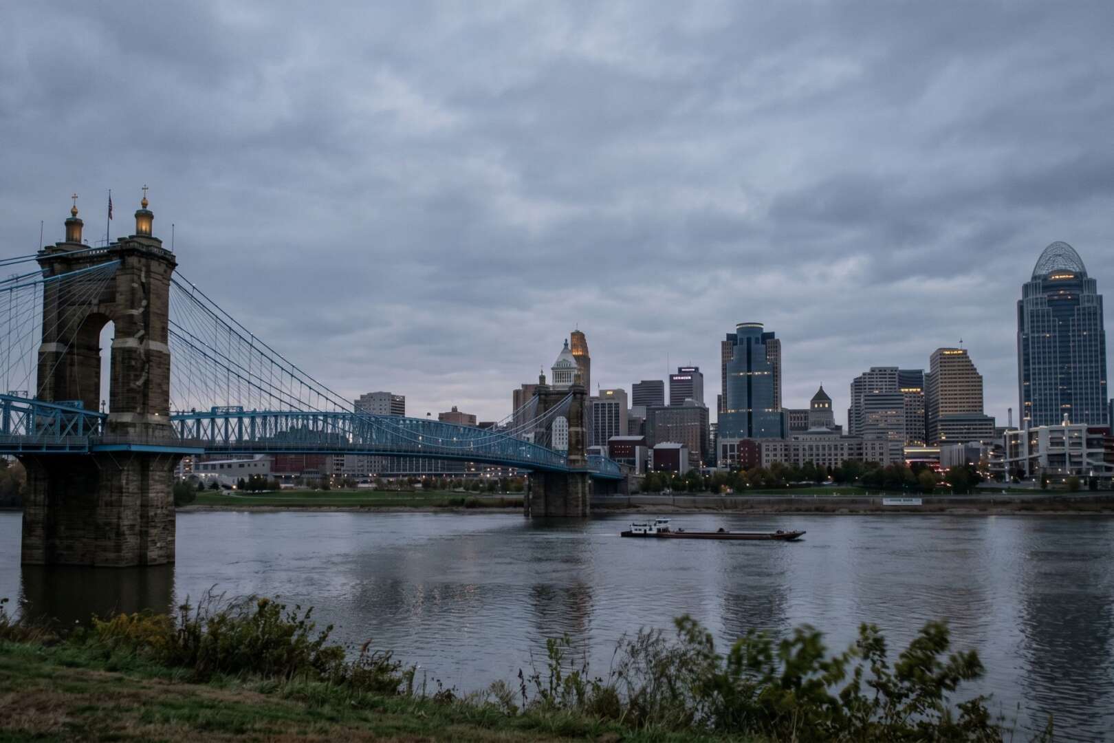 The Cincinnati skyline and John A. Roebling Suspension Bridge viewed across the Ohio River under an overcast sky.