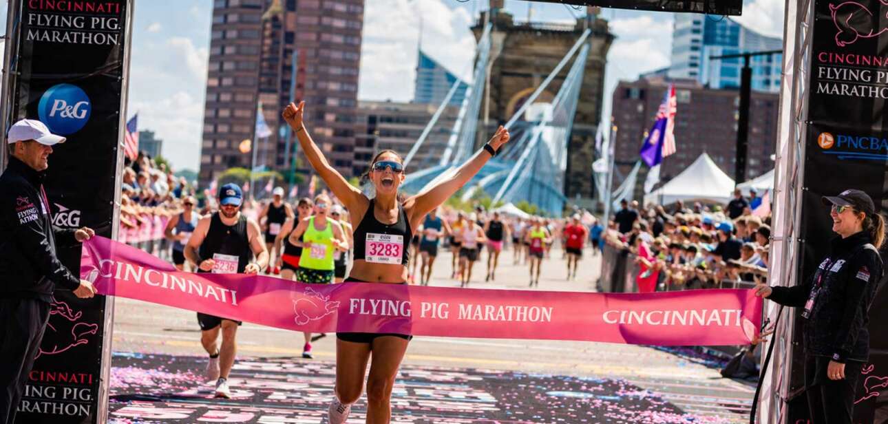 Finish line moment from the 2026 Flying Pig Marathon in Cincinnati, showing a runner completing the race with spectators, race officials, and the city skyline visible behind the course.