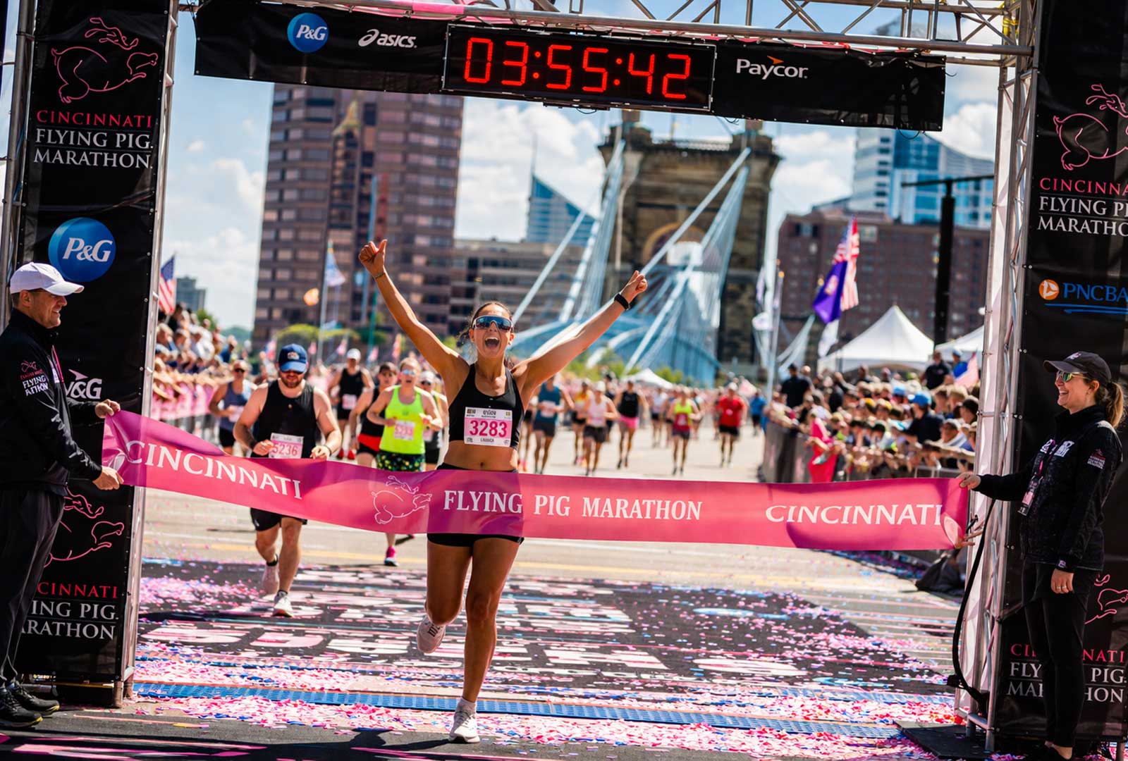 Finish line moment from the 2026 Flying Pig Marathon in Cincinnati, showing a runner completing the race with spectators, race officials, and the city skyline visible behind the course.