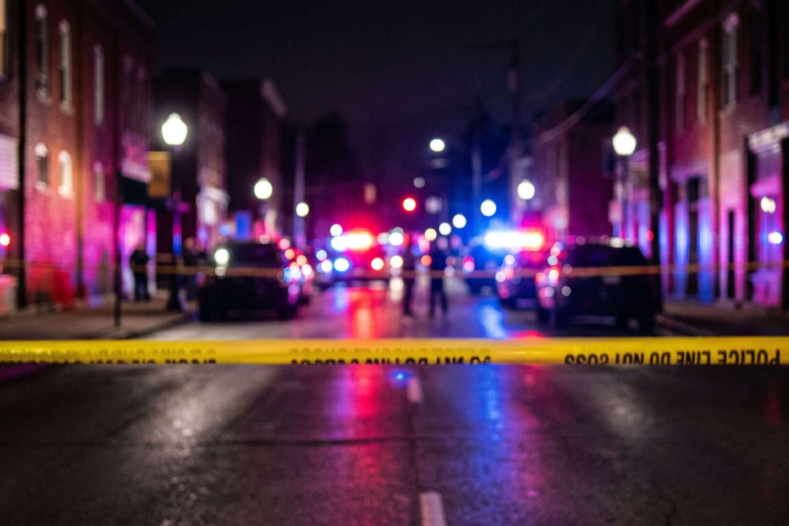Yellow police tape blocks a dark street with blurred, flashing emergency vehicle lights in the background at the scene of a fatal USPS delivery truck crash in St. Bernard.