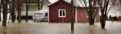 Deep floodwaters surrounding a dark red residential home and a recreational vehicle parked in the yard under a gloomy, overcast sky
