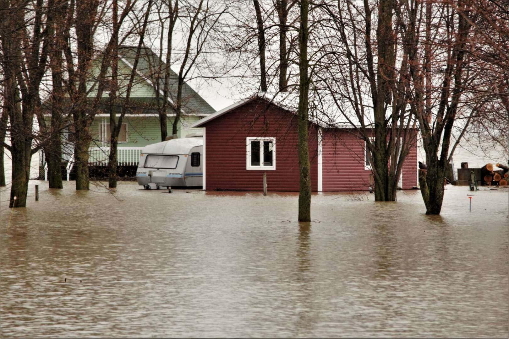 Deep floodwaters surrounding a dark red residential home and a recreational vehicle parked in the yard under a gloomy, overcast sky