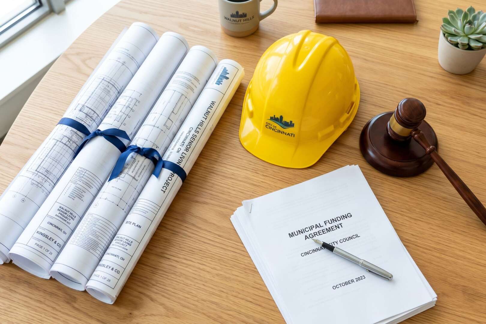 Close-up of a yellow hard hat, rolled blueprints, and a gavel on a wooden desk, representing the Kingsley housing grant project in Walnut Hills, Cincinnati.