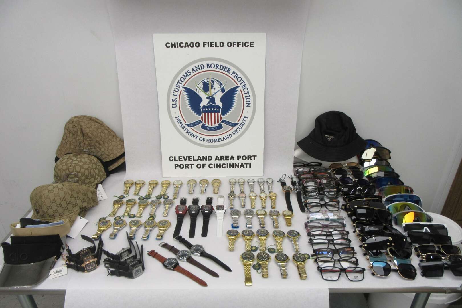 A display table showing various seized counterfeit designer accessories, including watches and sunglasses, with a U.S. Customs and Border Protection seal in the background.