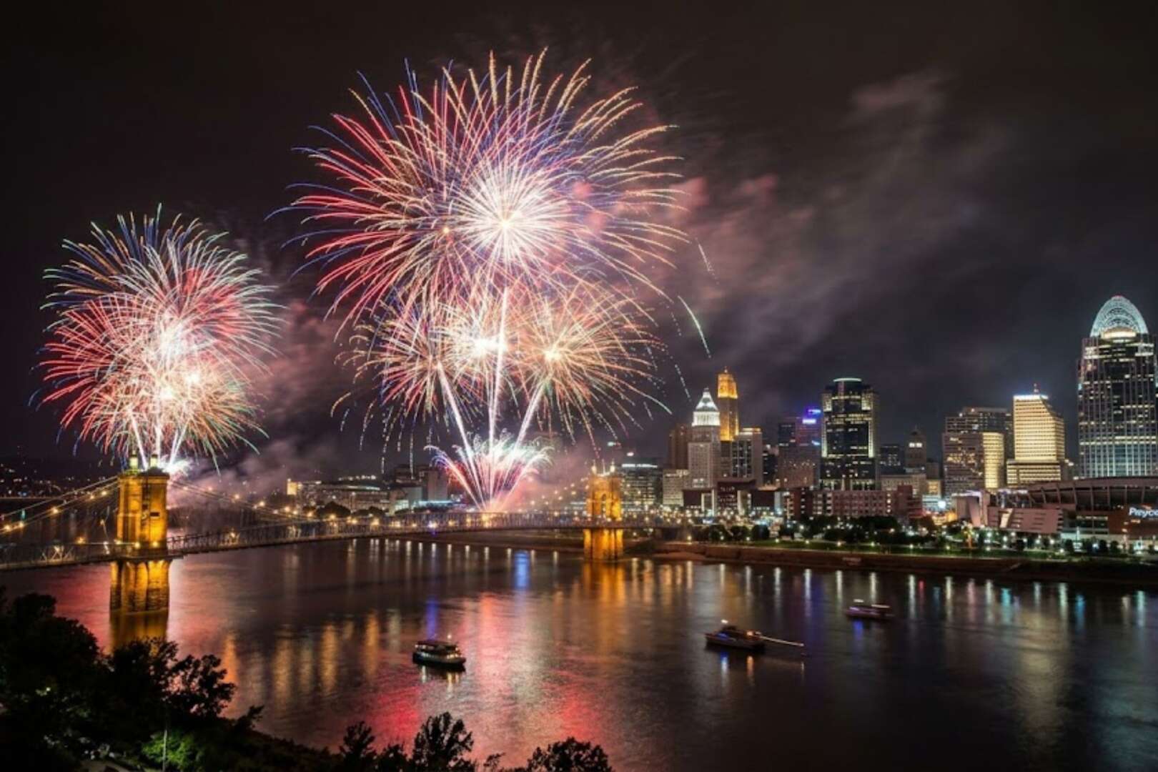 Bright fireworks exploding over the Cincinnati city skyline and the John A. Roebling Suspension Bridge at night.