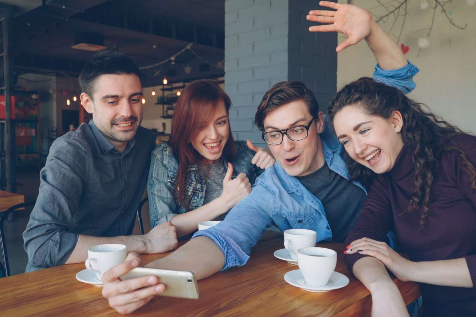 Four young Gen Z adults smiling and looking at a smartphone together while sitting at a table in a cafe.
