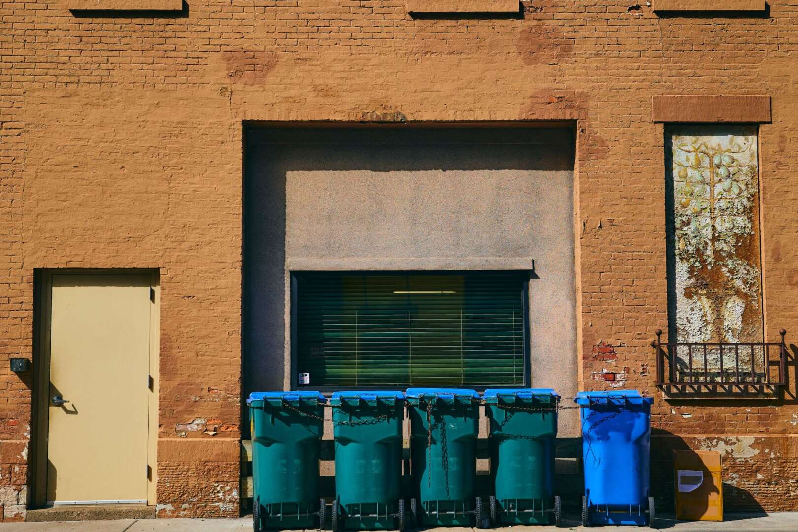 A row of four waste bins, three green and one blue, lined up against an urban brick building in Cincinnati.