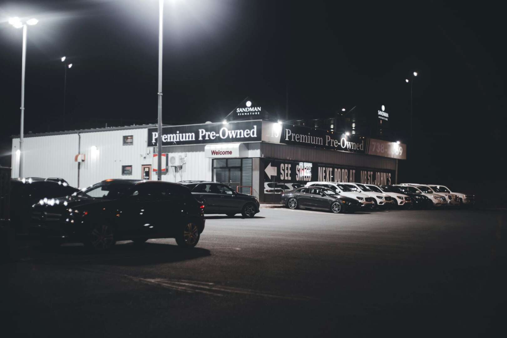 A nighttime view of a 'Premium Pre-Owned' used car dealership lot with vehicles parked under bright lights.