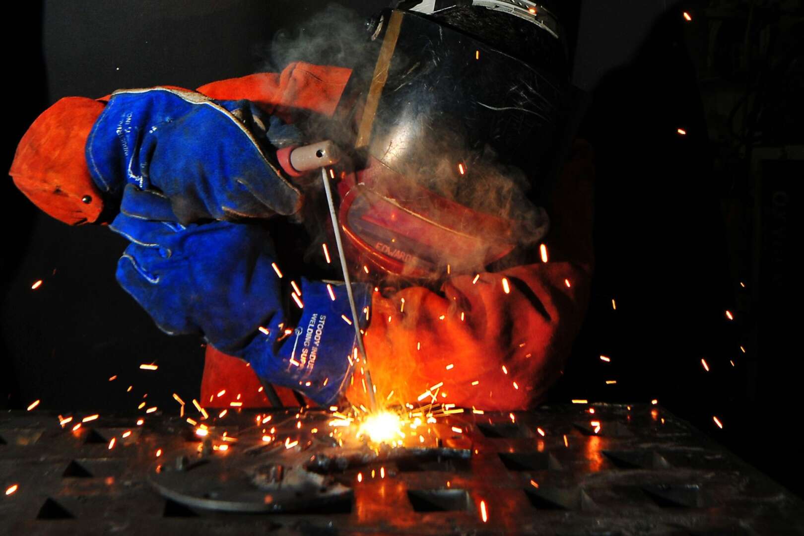 An industrial worker wearing a blue protective suit and helmet welding metal with bright orange sparks flying.