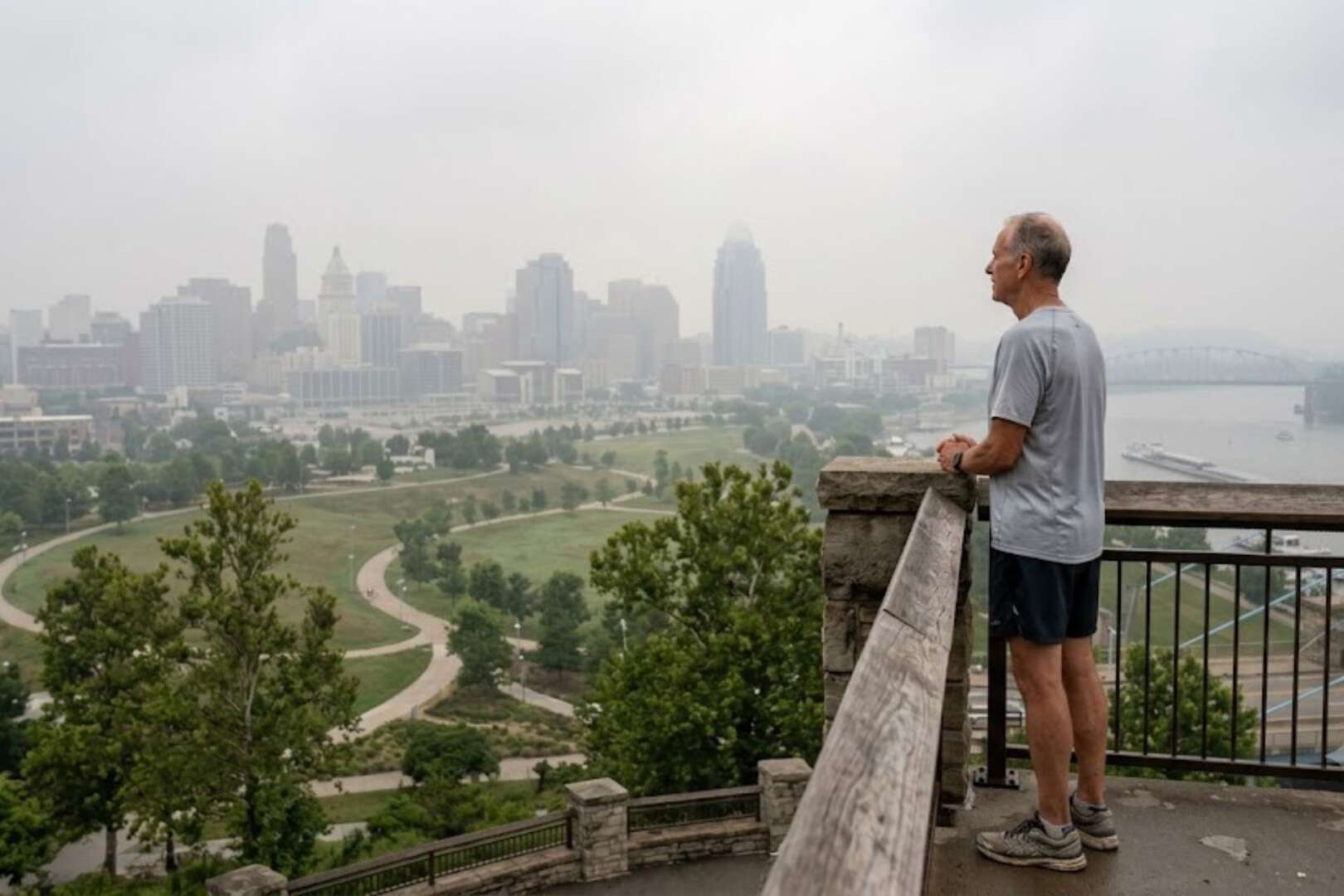 A man stands on a scenic overlook looking out at the Cincinnati city skyline, which is heavily obscured by thick smog and poor air quality.