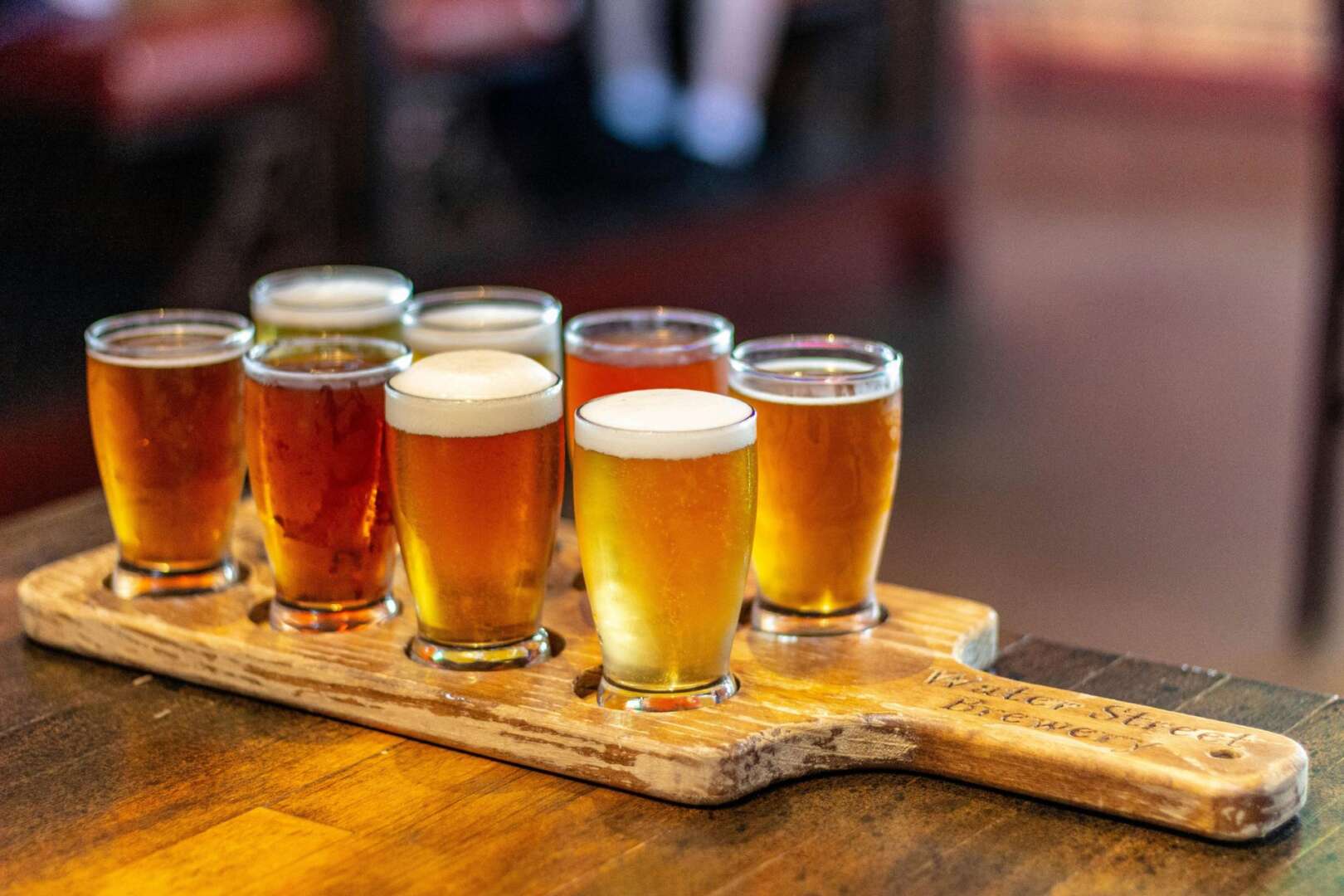 A row of six craft beer samples in taster glasses served on a wooden flight board.