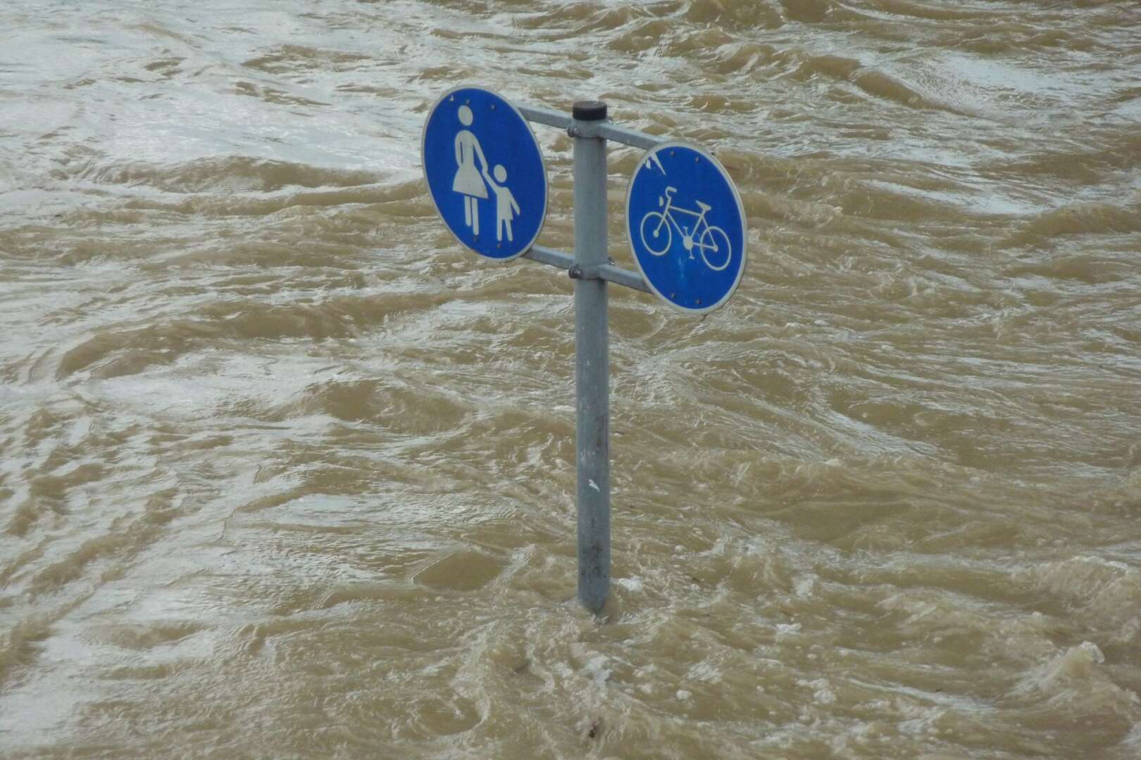 A blue pedestrian and bicycle path sign partially submerged in deep, muddy floodwater.