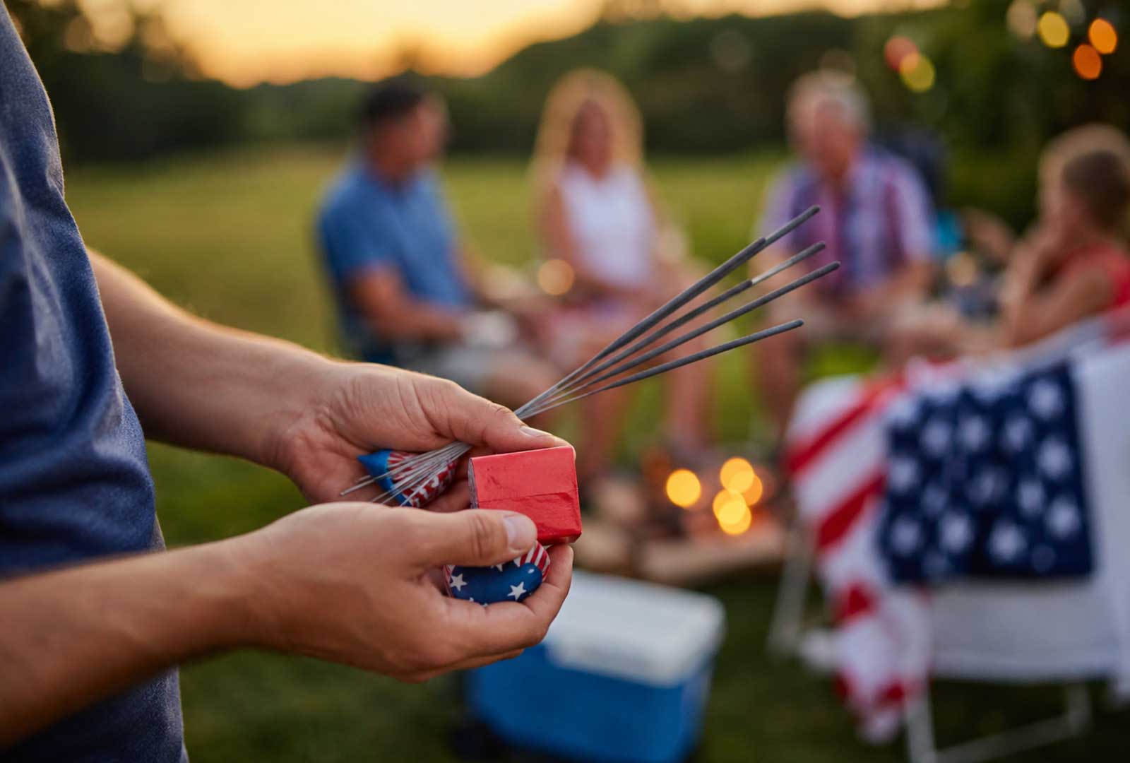 Adult hands holding unlit sparklers and small fireworks during a Fourth of July gathering, with family blurred in the background.