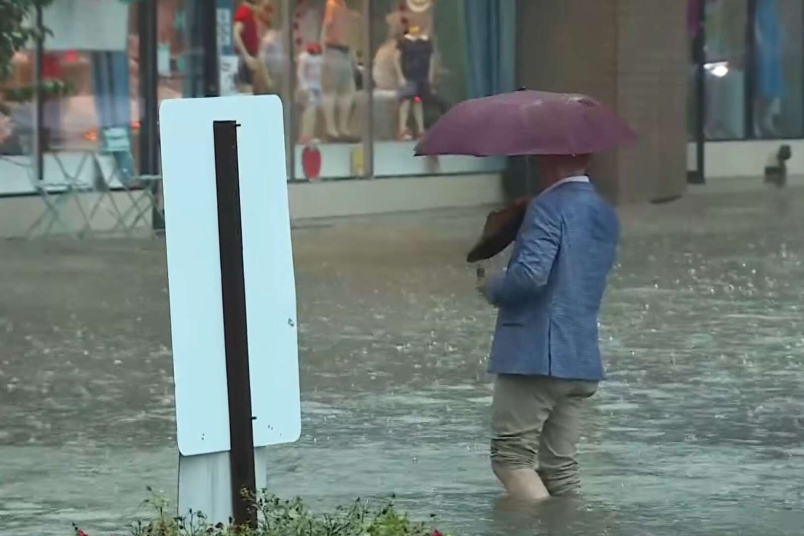 A person holding an umbrella wades through knee-deep floodwaters on a city street in front of a storefront.