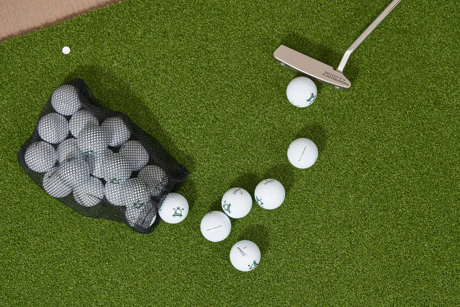 Overhead view of a mini golf putter and scattered white golf balls on green artificial turf next to a basket of golf balls.