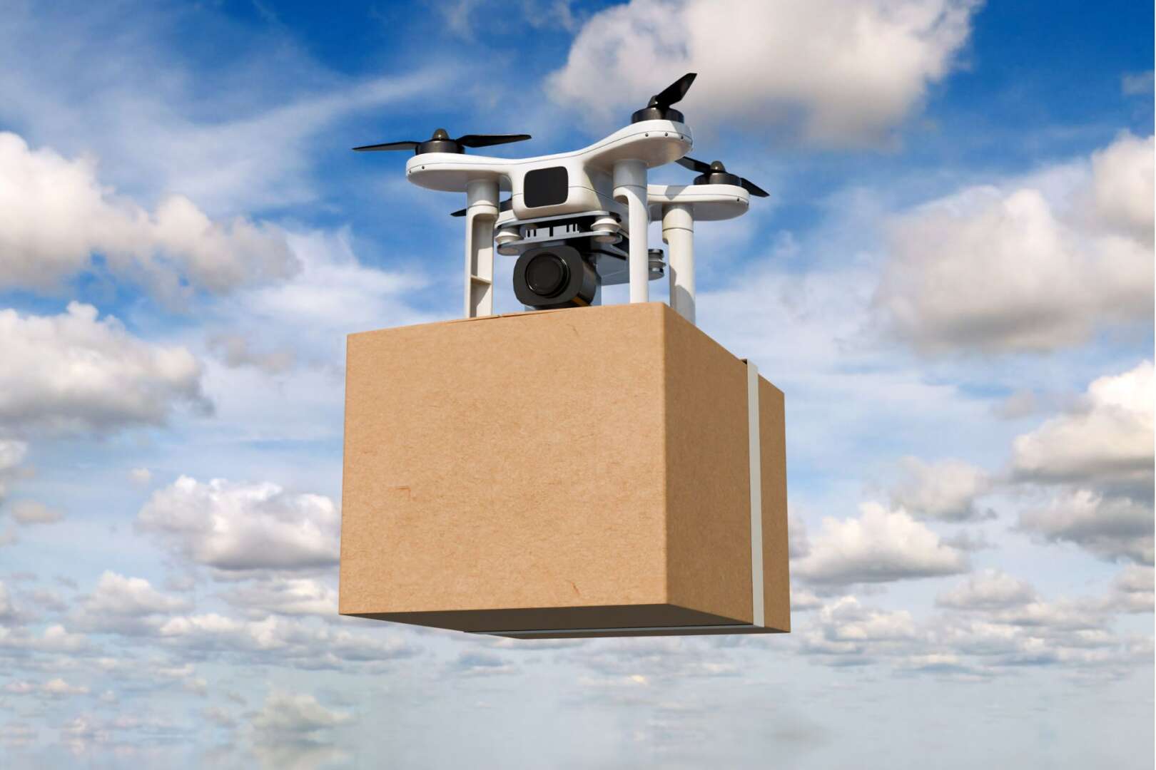 A white quadcopter drone in mid-flight carrying a cardboard delivery package against a cloudy blue sky.