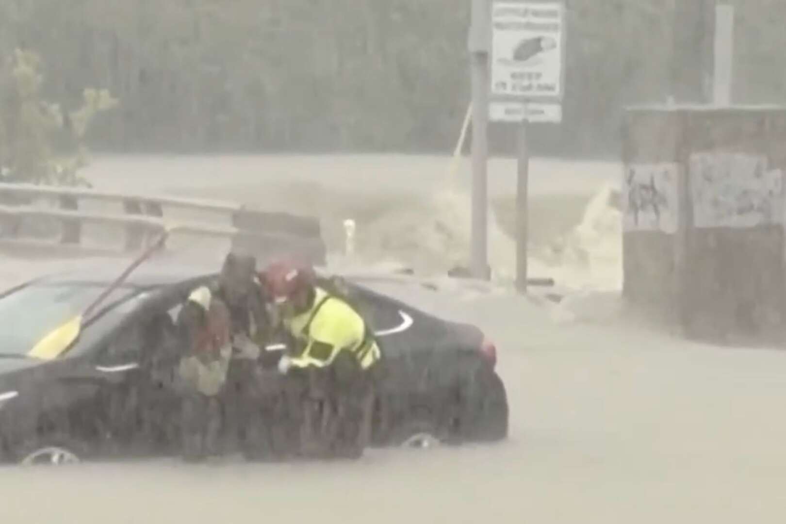 First responders in yellow high-visibility jackets and helmets wading through deep floodwaters to reach a partially submerged black car.