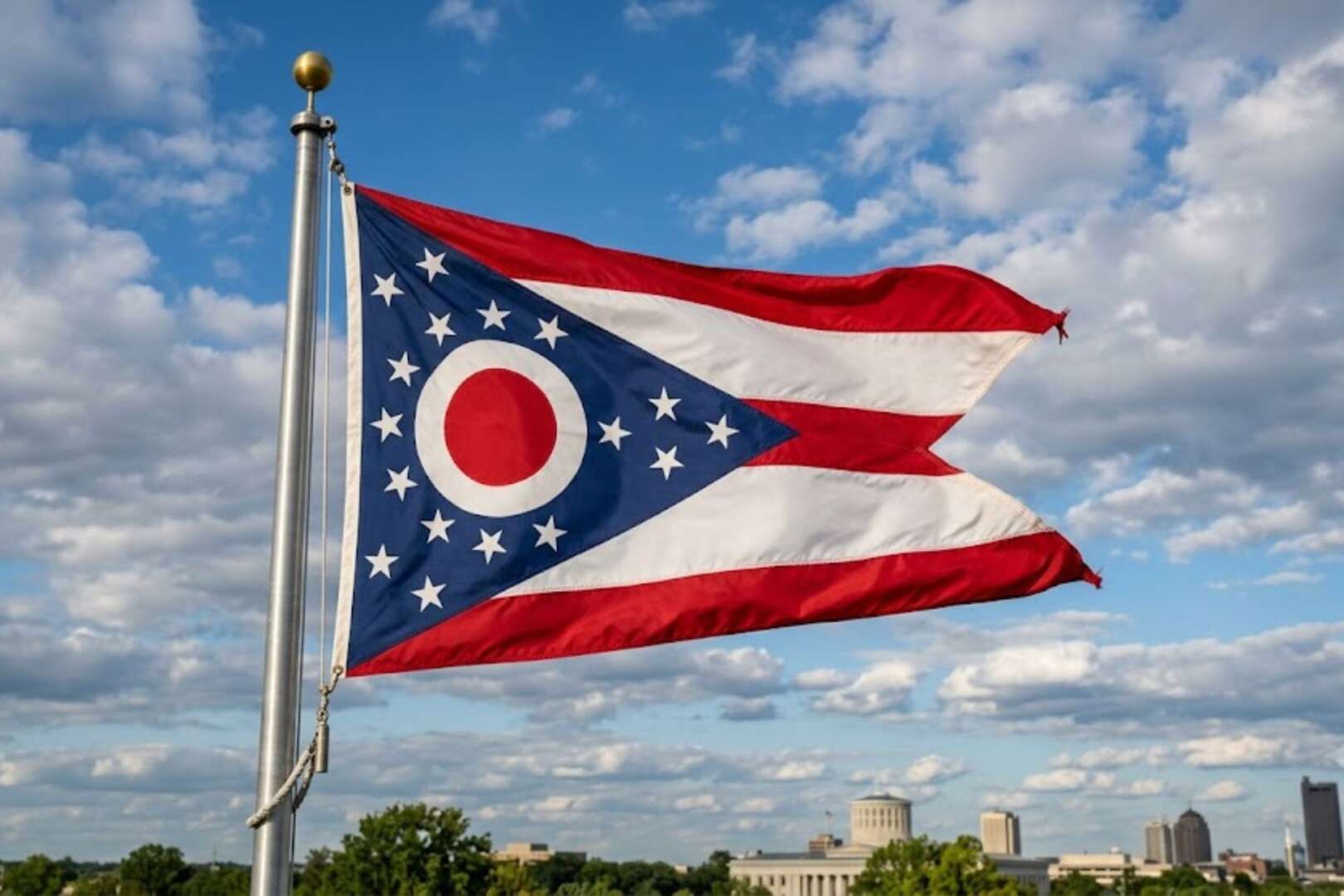 The state flag of Ohio waving on a flagpole against a partly cloudy sky with a city skyline in the background.