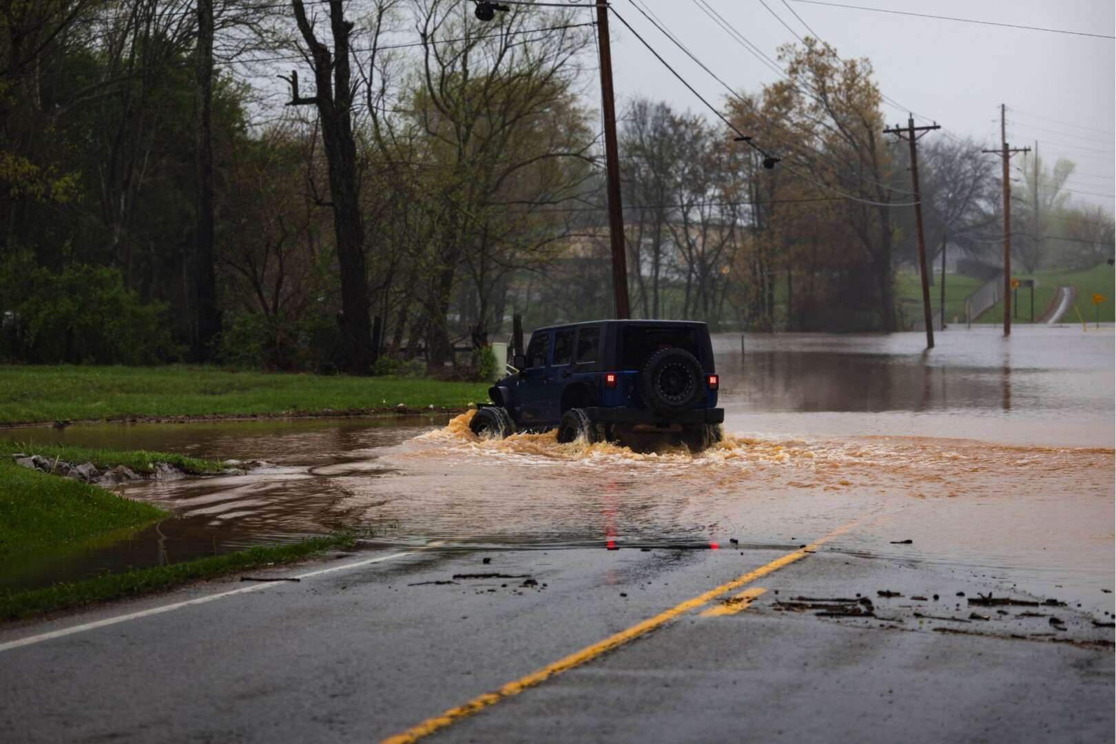 A jeep traverses a flooded road surrounded by trees, showcasing a challenging weather condition.
