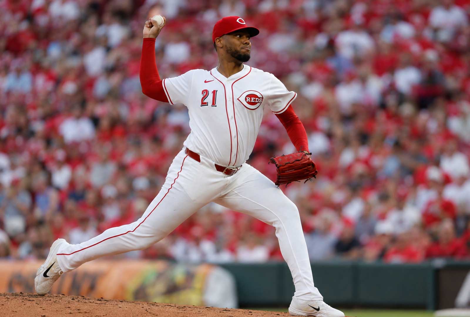 Photorealistic image of a Cincinnati Reds pitcher throwing from the mound at Great American Ball Park with a blurred red crowd behind him.