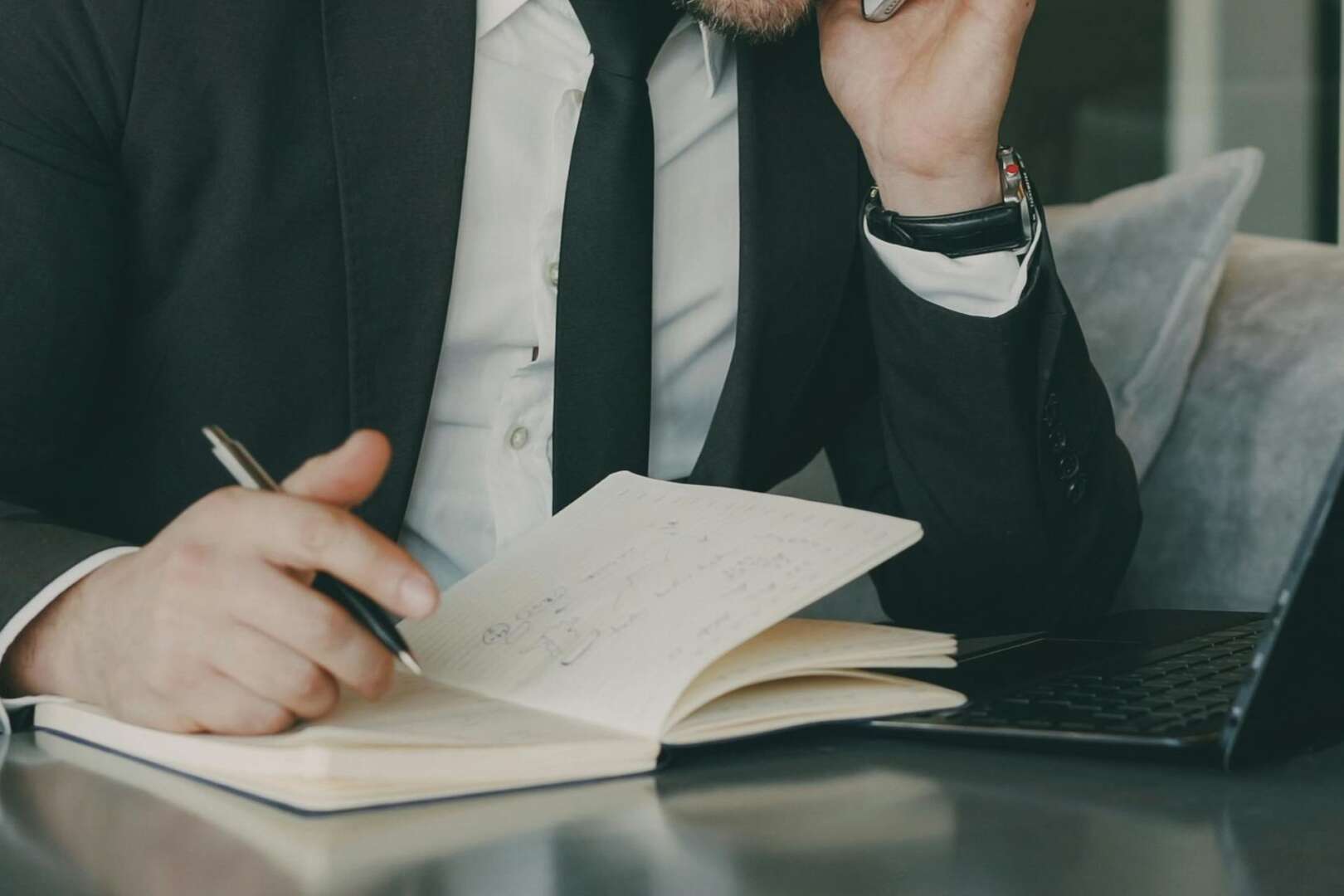 A businessman in a dark suit holding a pen over a notebook at a desk, appearing stressed or deep in thought.