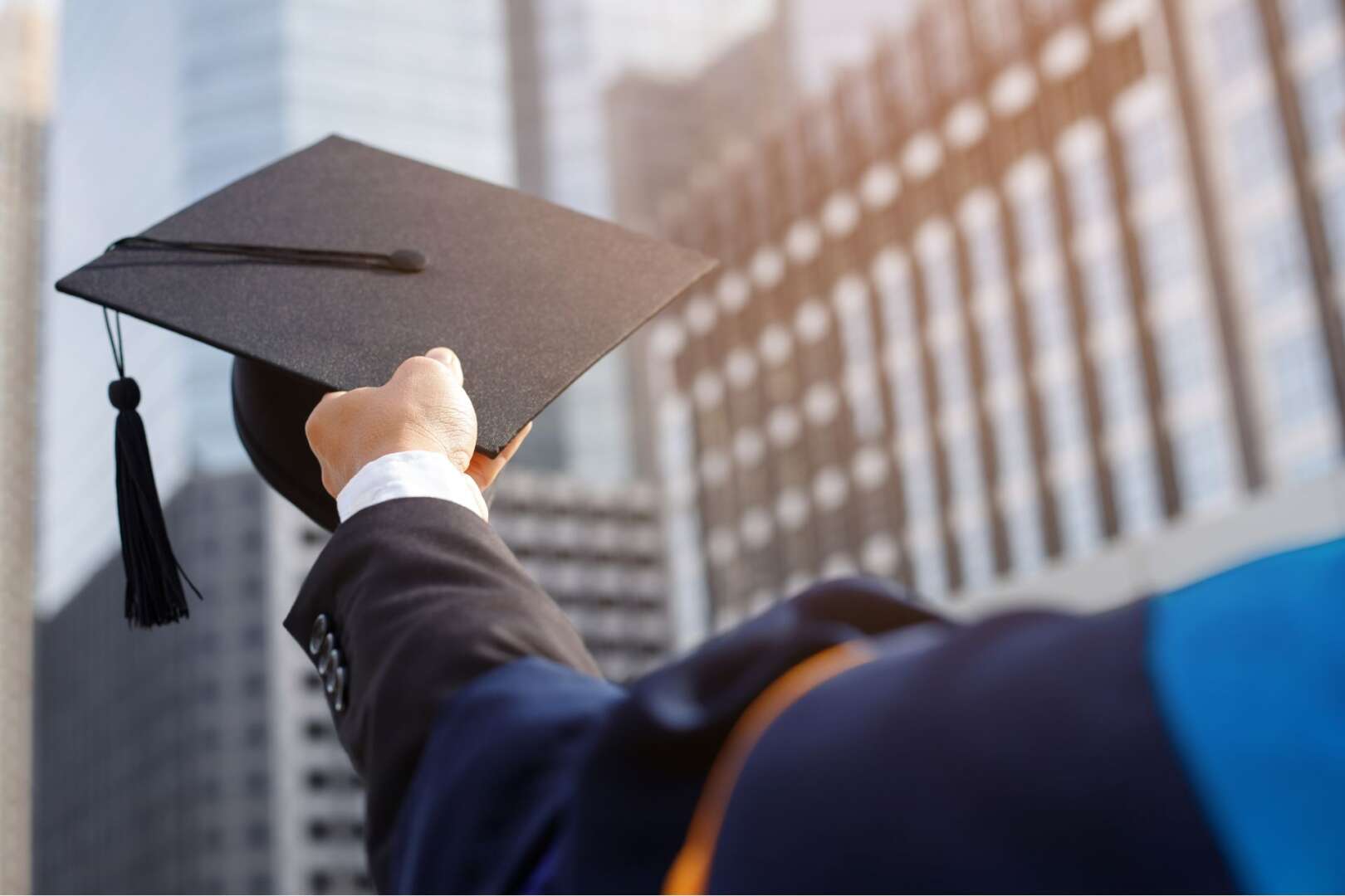 A graduate holding up a graduation cap against a backdrop of modern campus buildings.