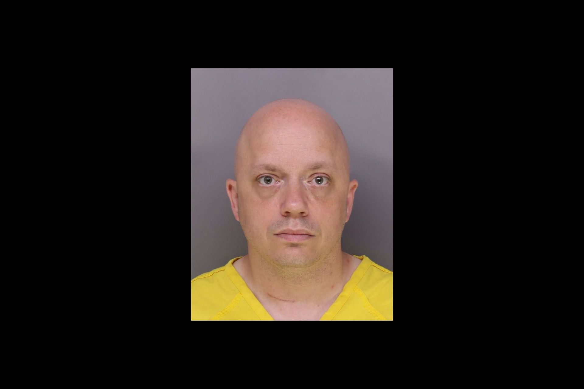 Mugshot photo of a bald male wearing a yellow shirt against a dark background.