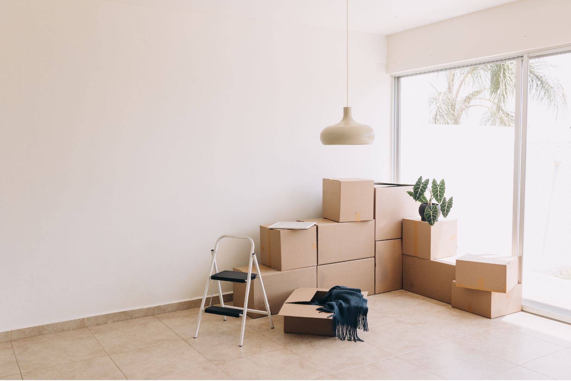 Cardboard moving boxes and a step stool stacked tightly in a brightly lit, minimalist apartment with light wood floors.