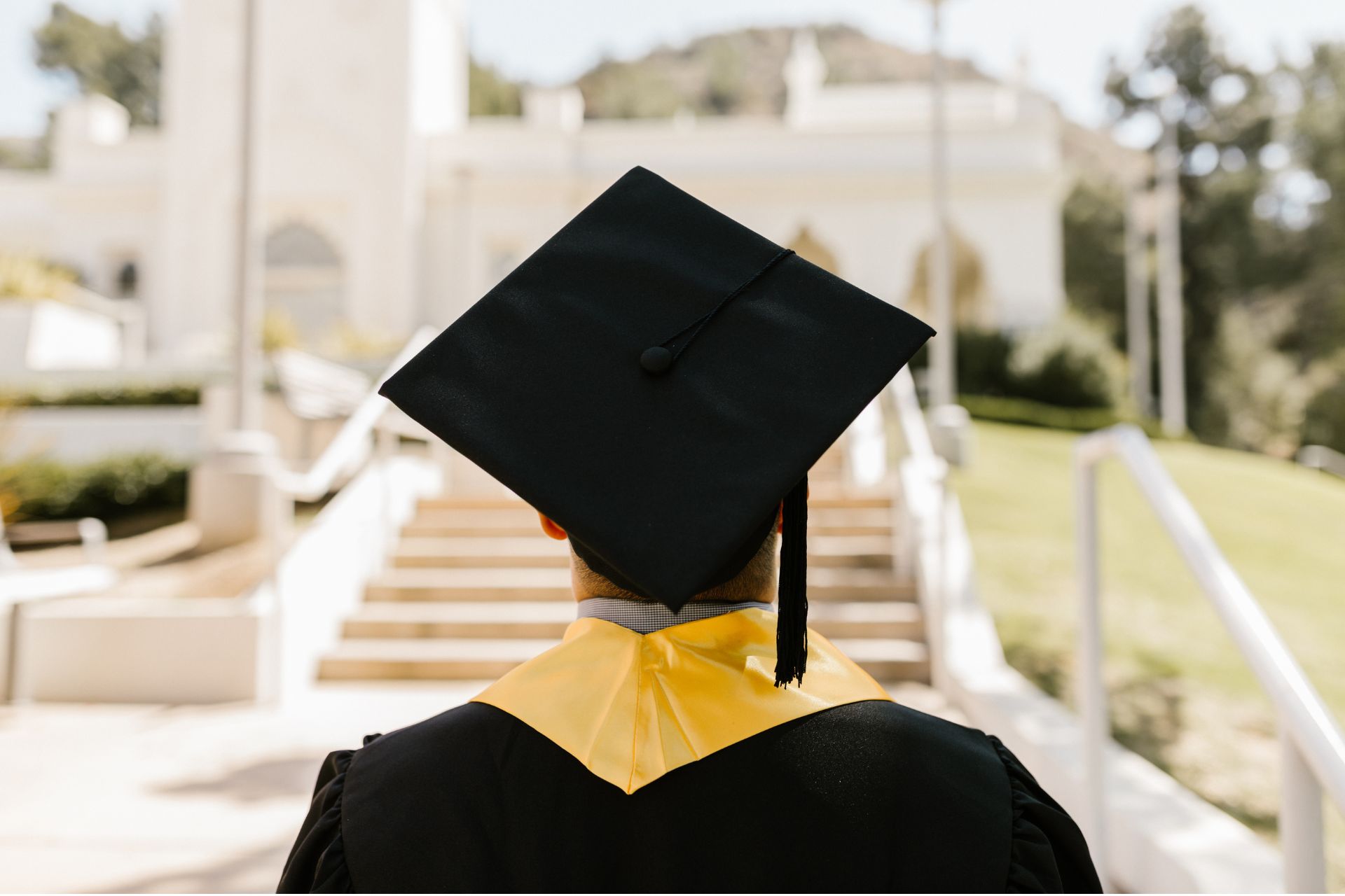 Rear view of a university graduate wearing a black graduation cap and gown with a yellow sash, walking up outdoor steps toward a campus building.