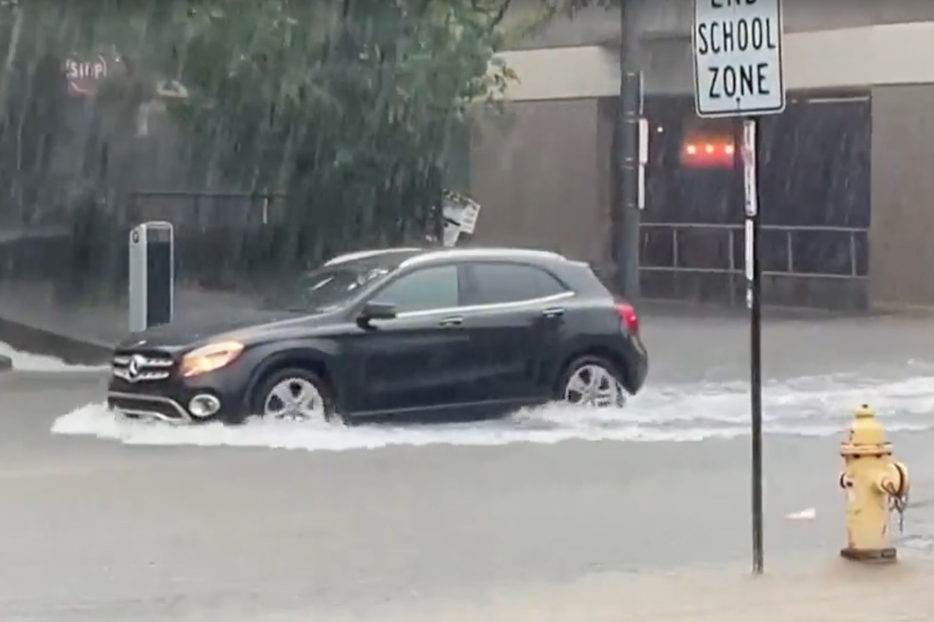 Black car driving through deep floodwaters on a city street during heavy rain.