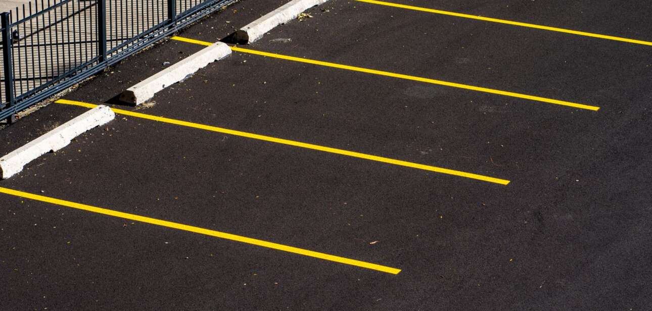 Empty outdoor parking lot with yellow painted stall lines and white wheel stops along a perimeter fence.