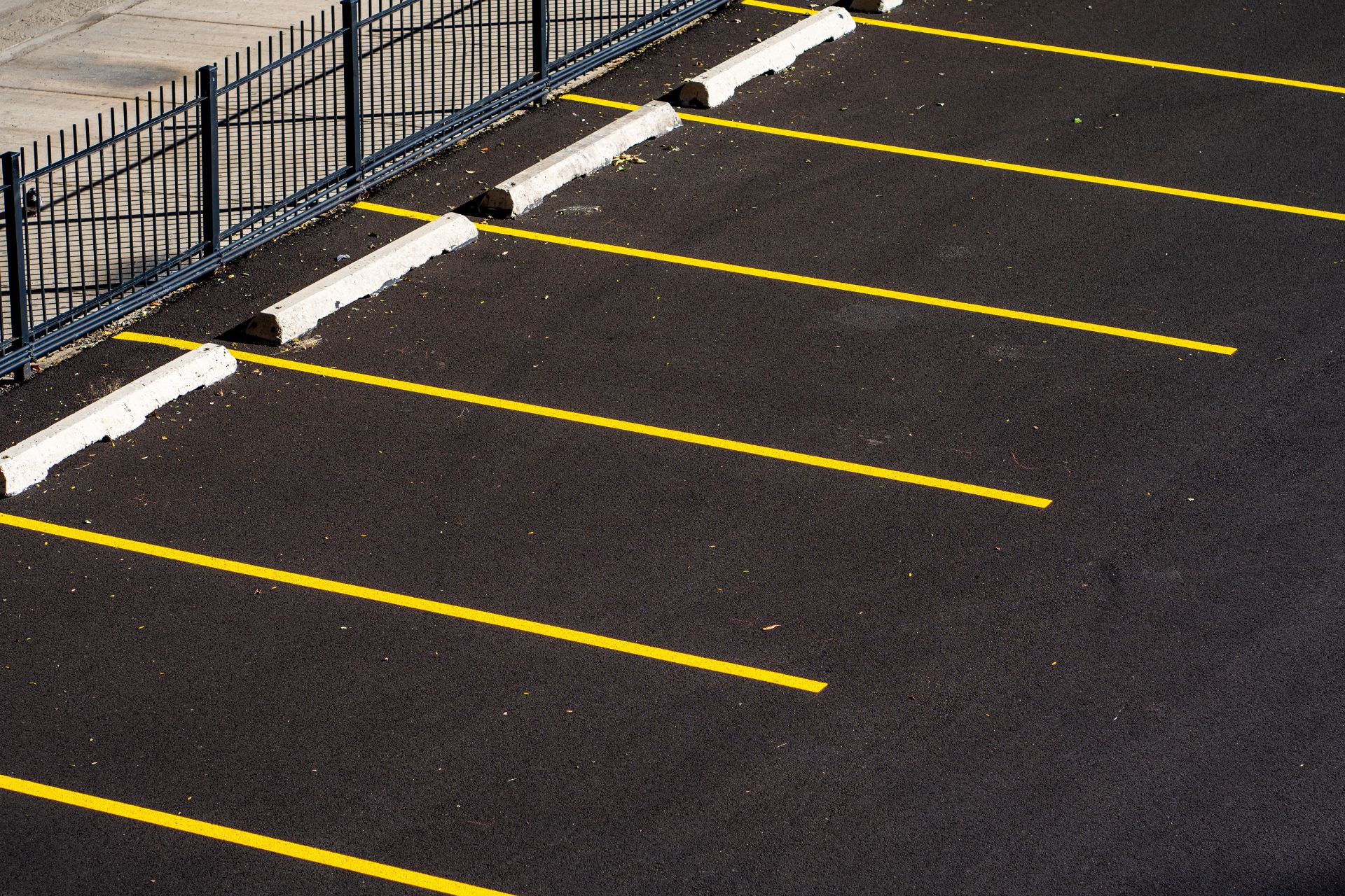 Empty outdoor parking lot with yellow painted stall lines and white wheel stops along a perimeter fence.