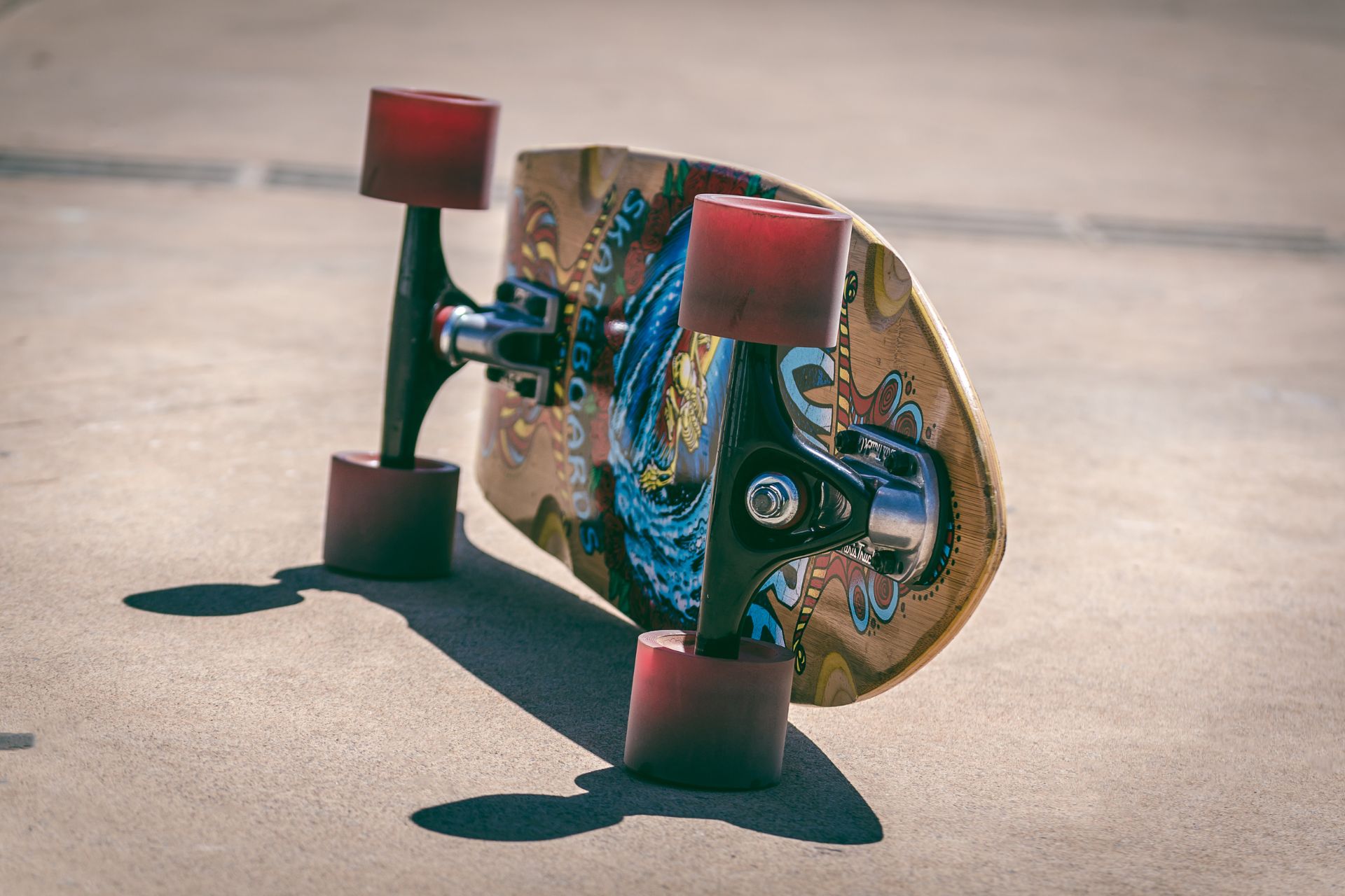 An inverted wooden skateboard resting on concrete, exposing red wheels, metallic trucks, and an intricate undersurface graphic design.
