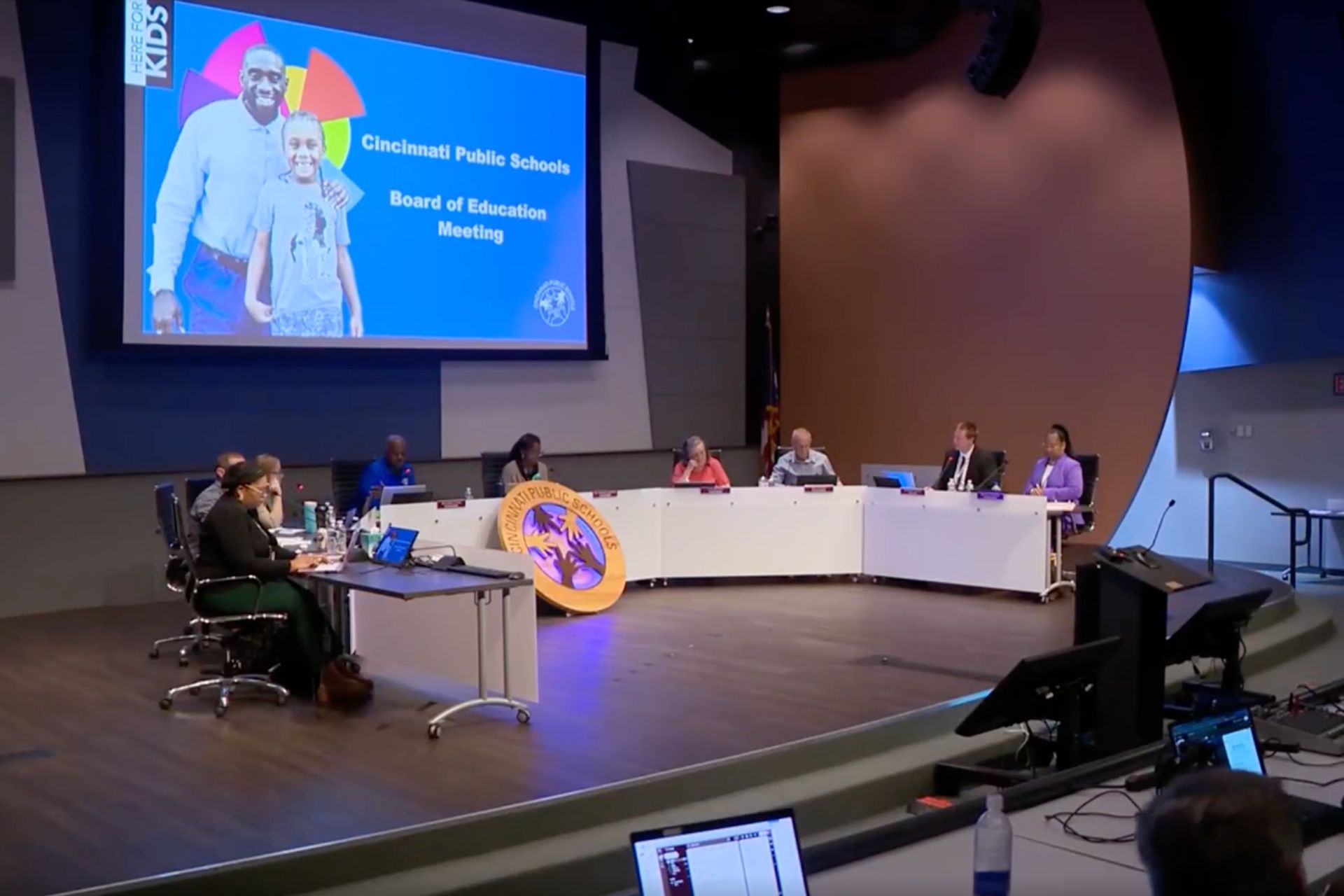 Cincinnati Public Schools board members seated at a U-shaped desk during a meeting in an auditorium, with a presentation screen displaying in the background.