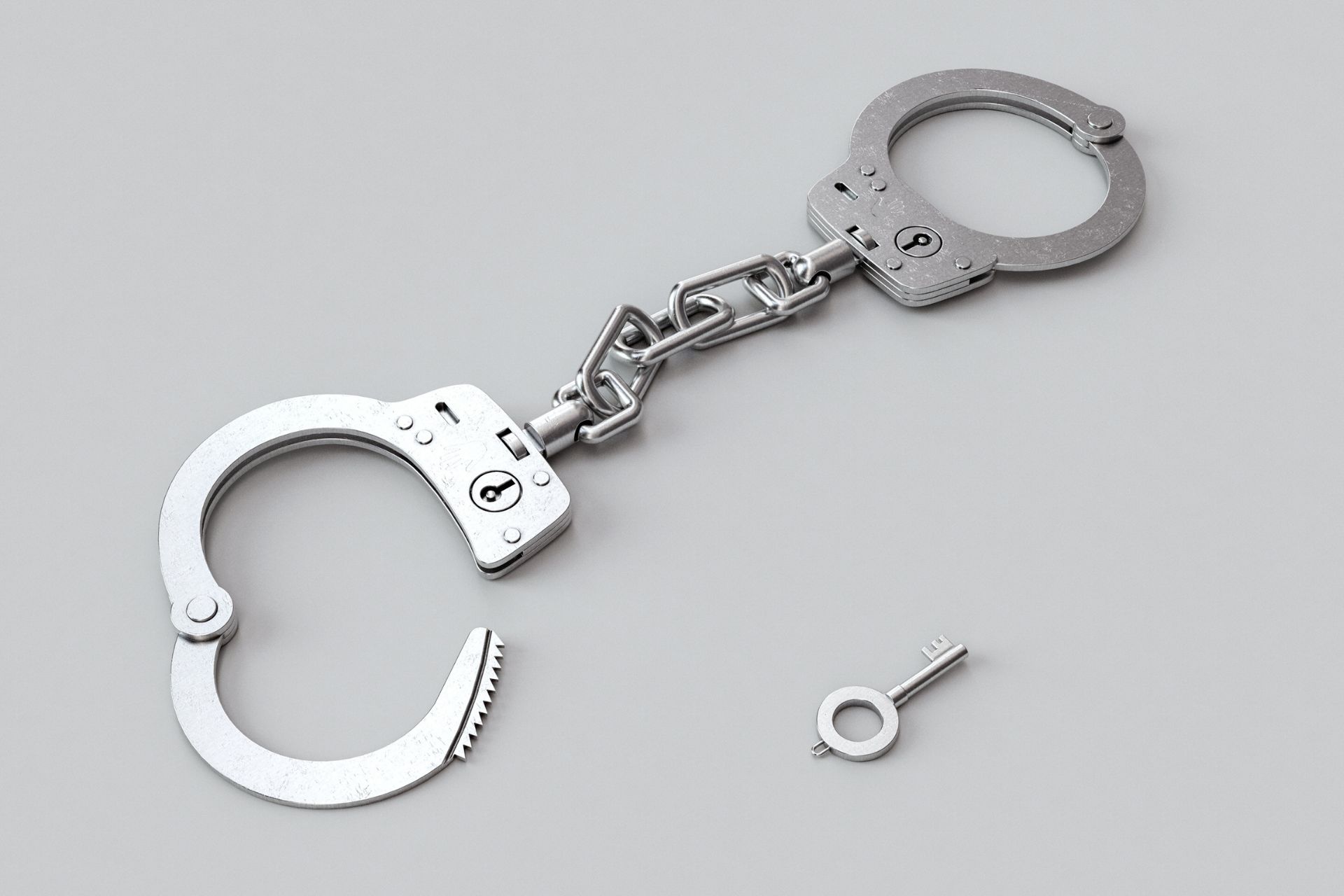 Pair of open silver metal handcuffs and a small key lying on a plain gray surface.