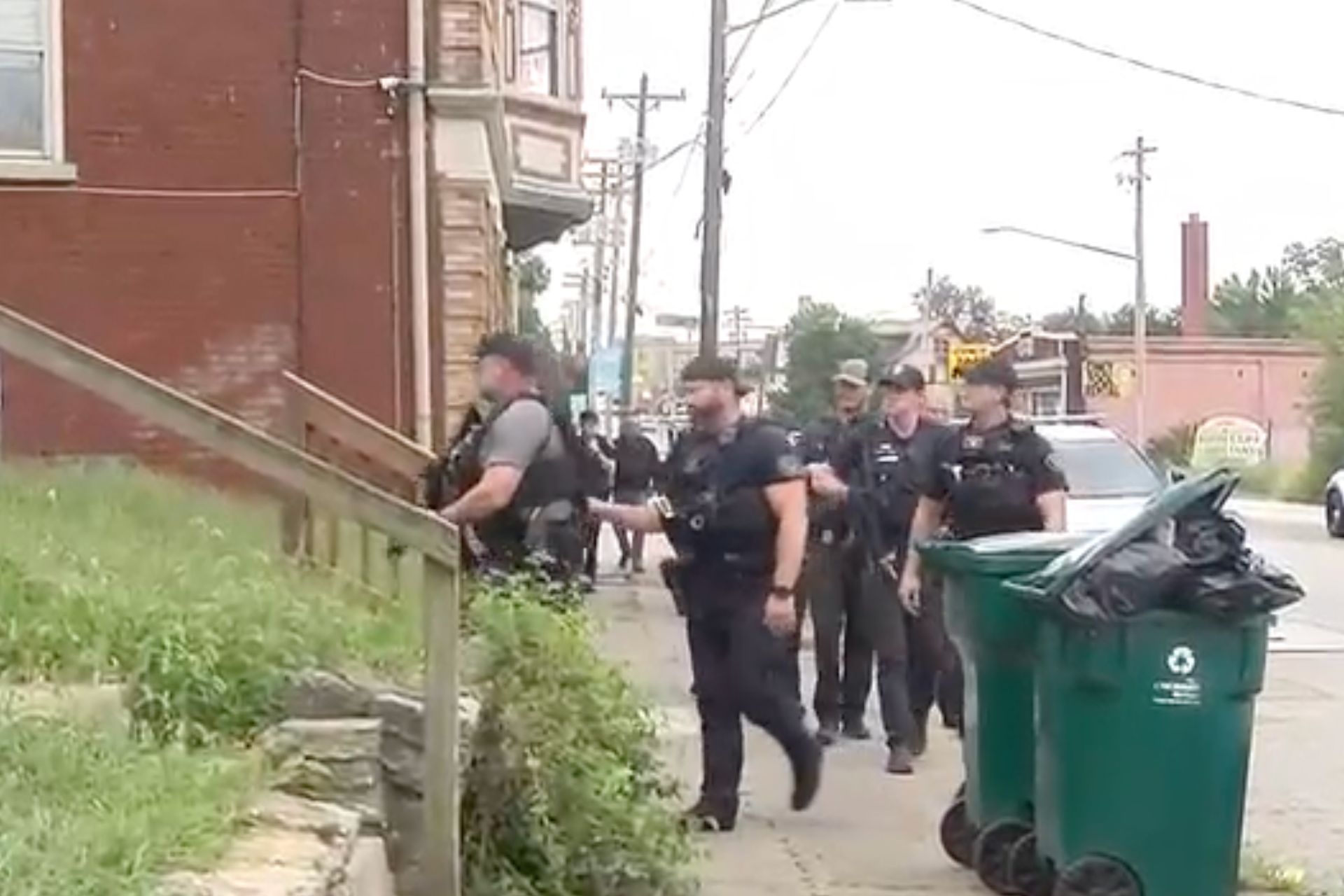 Several Cincinnati police officers walking down a residential sidewalk past a brick building and green trash bins.