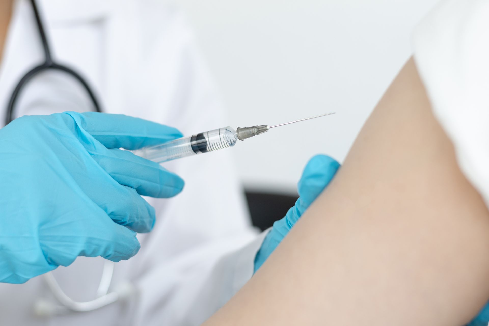 A healthcare professional in a white coat and blue gloved hands preparing a syringe to administer a vaccine into a patient's upper arm.