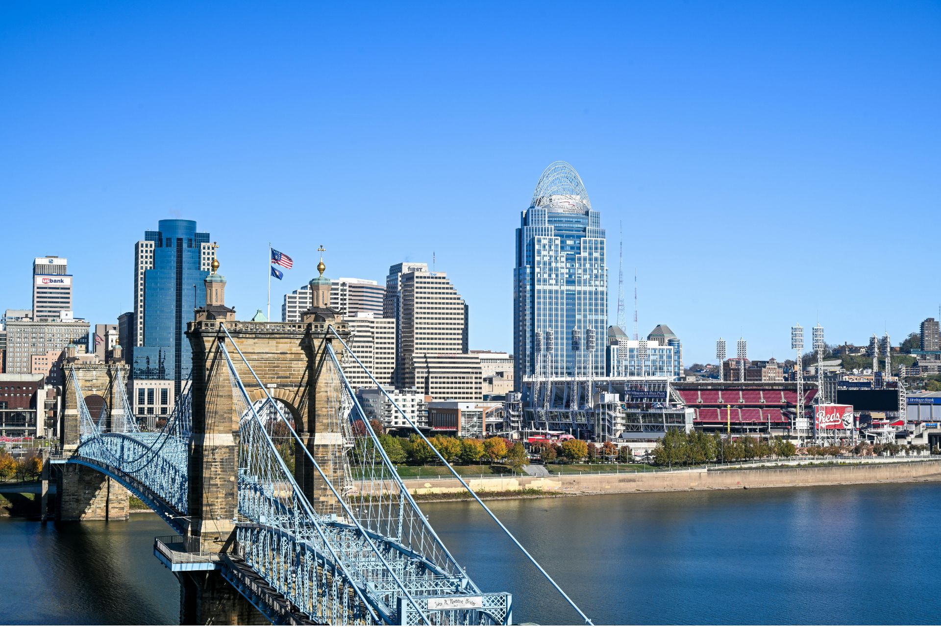 John A. Roebling Suspension Bridge leading toward the Downtown Cincinnati skyline across the Ohio River under a clear blue sky.