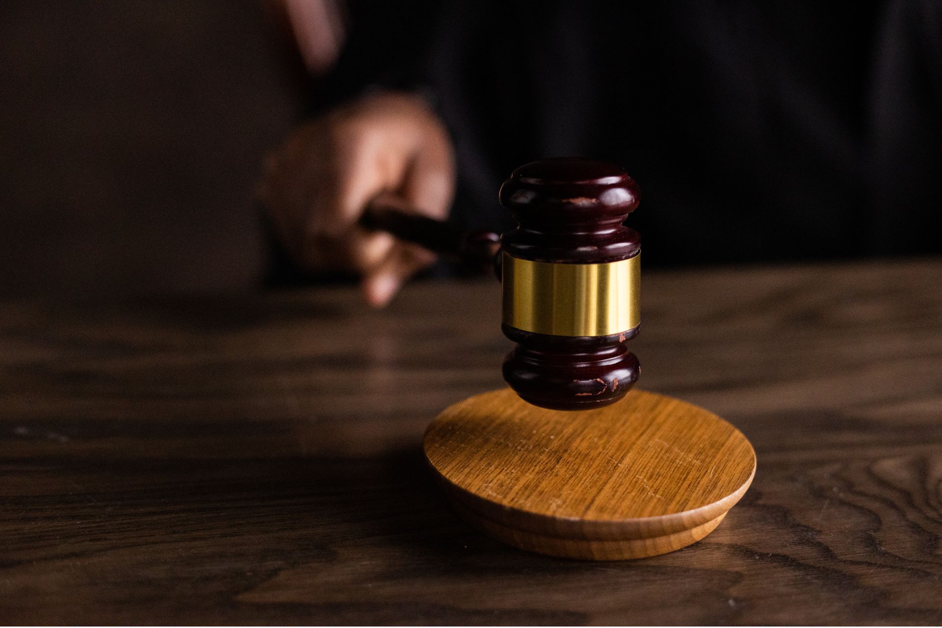 A hand holding a wooden judge's gavel resting on a sound block on a dark wooden desk.
