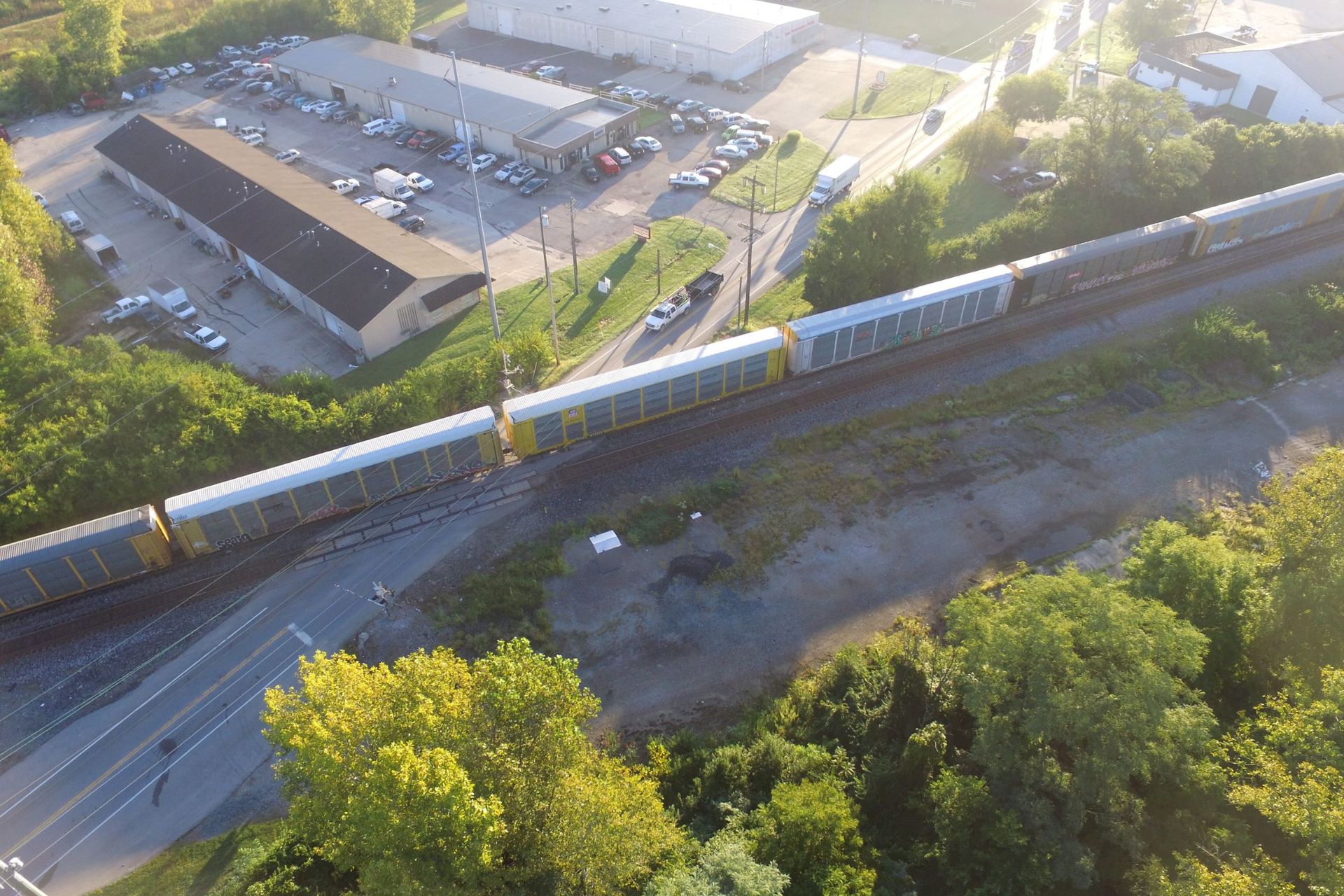 High-angle aerial photograph of a freight train traveling on curved railroad tracks surrounded by lush trees and industrial buildings.