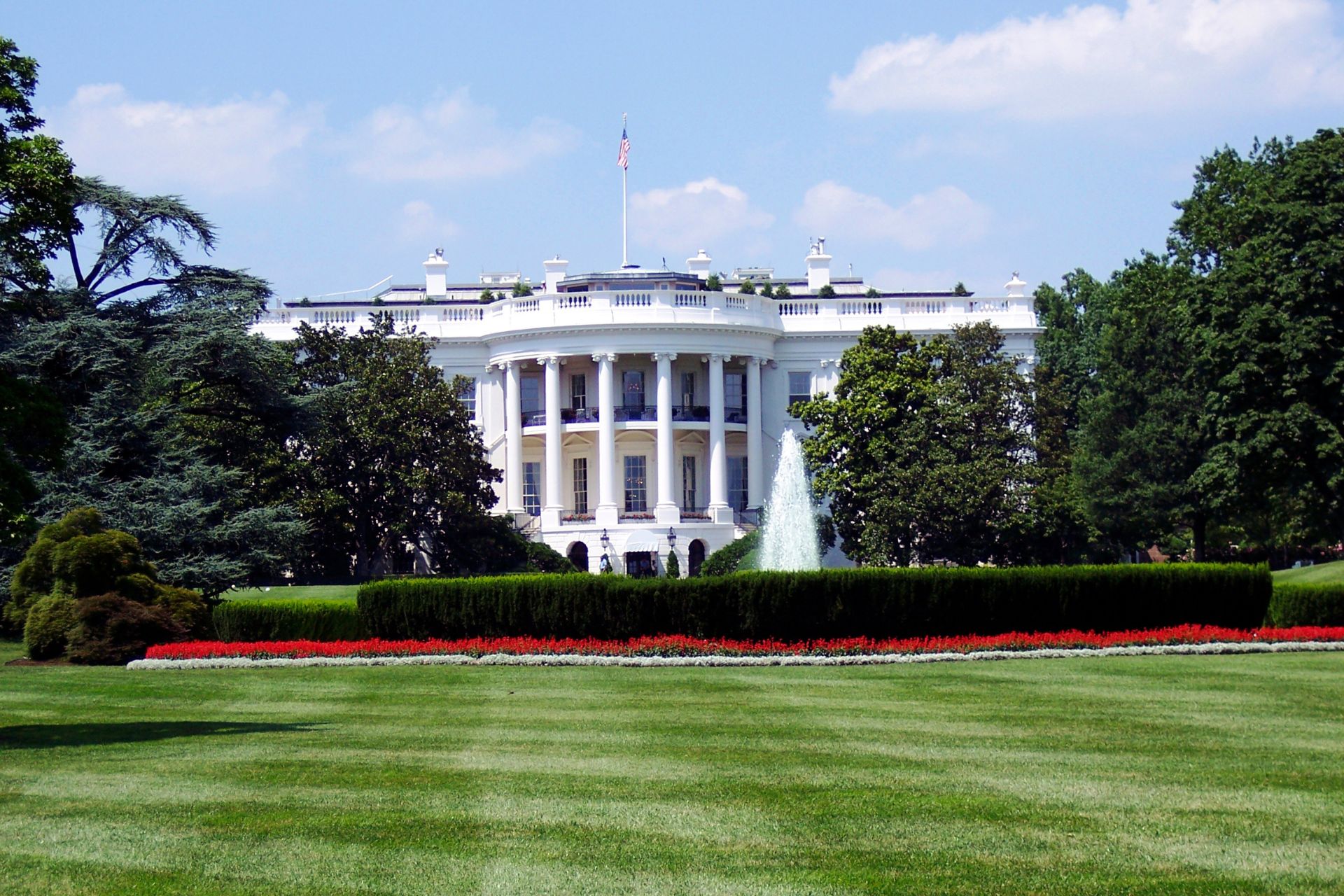 Exterior view of the White House framed by a green lawn, trees, and a clear blue sky with light clouds.