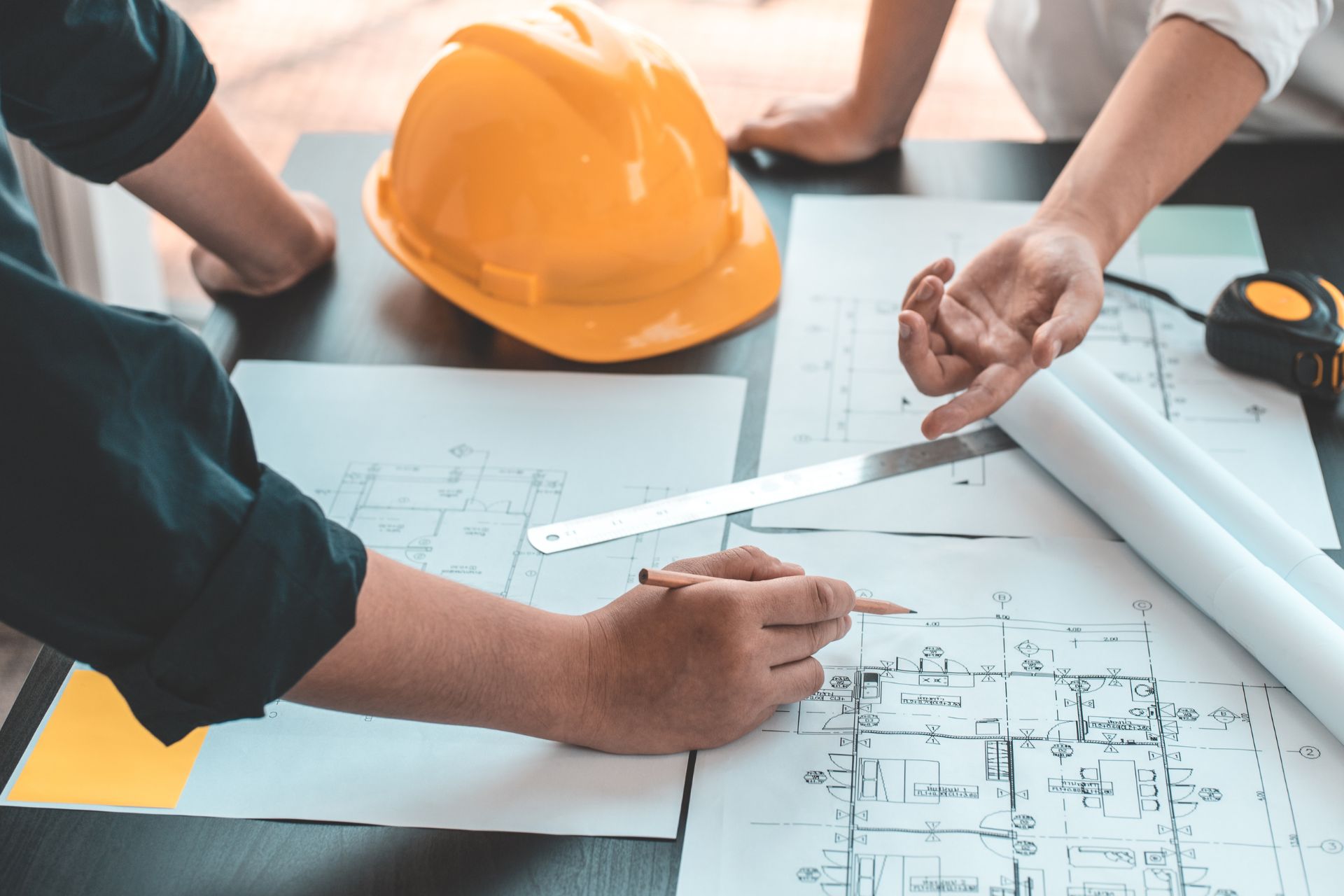 Construction professionals and engineers pointing at blueprints on a table with a yellow hard hat and measuring tape.