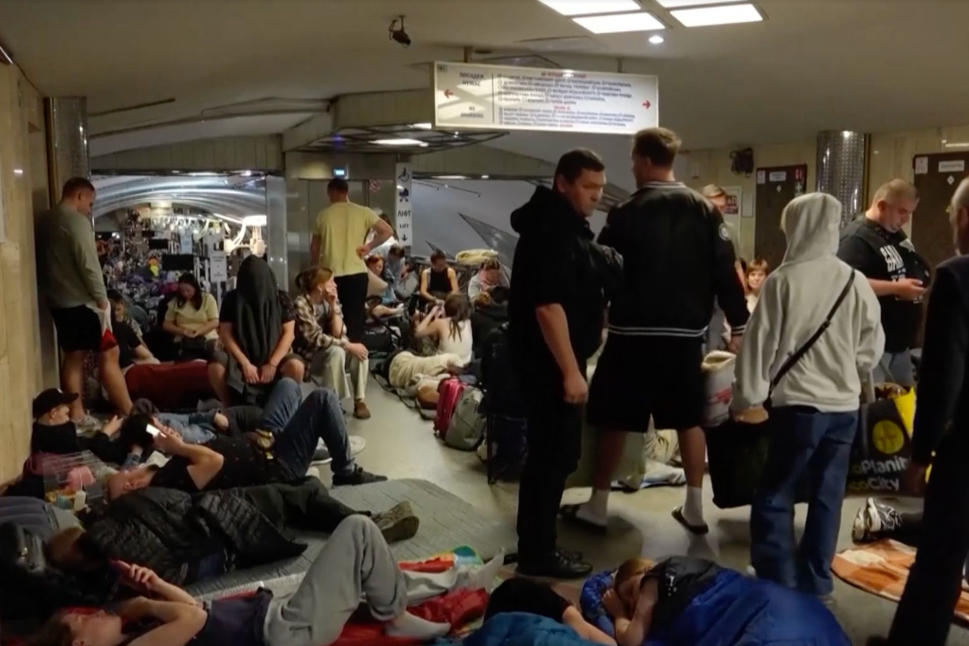 People gathered in a crowded indoor facility with mats and belongings spread across the floor.