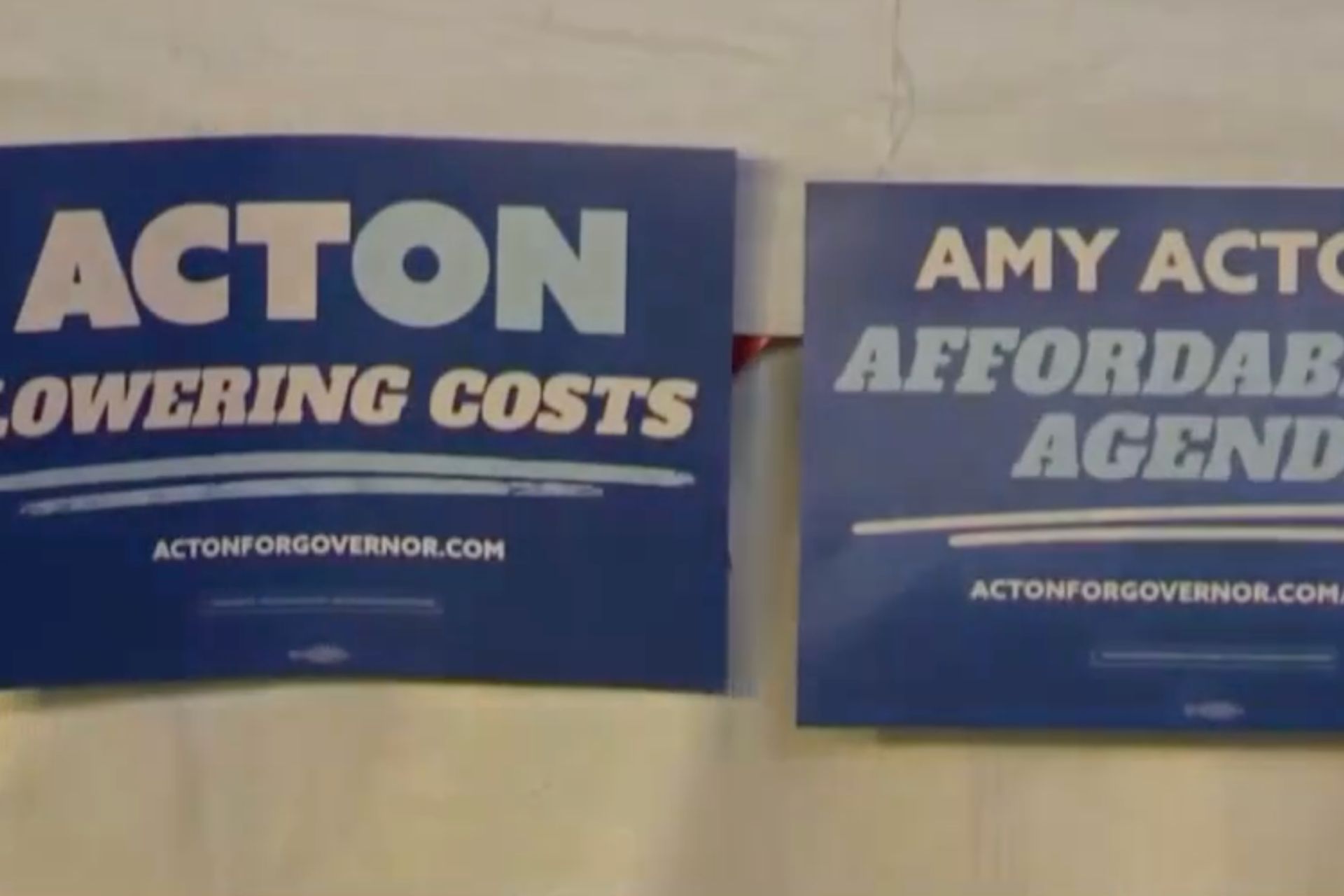 Two blue Amy Acton for Governor campaign signs displaying the text "Acton Lowering Costs" and "Amy Acton Affordable Agenda."