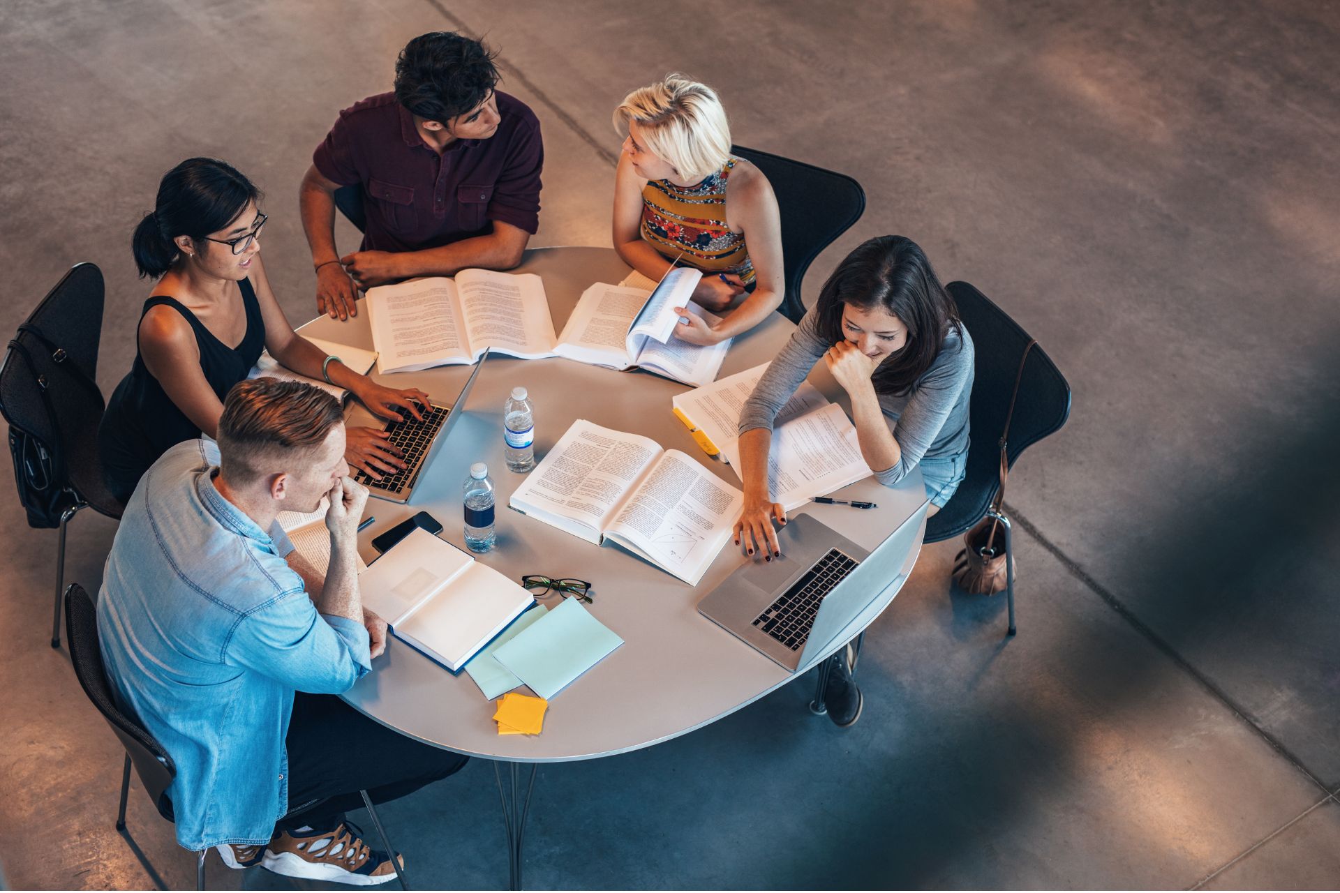 High-angle view of a group of college students sitting around a round table studying together with open books and notebooks.