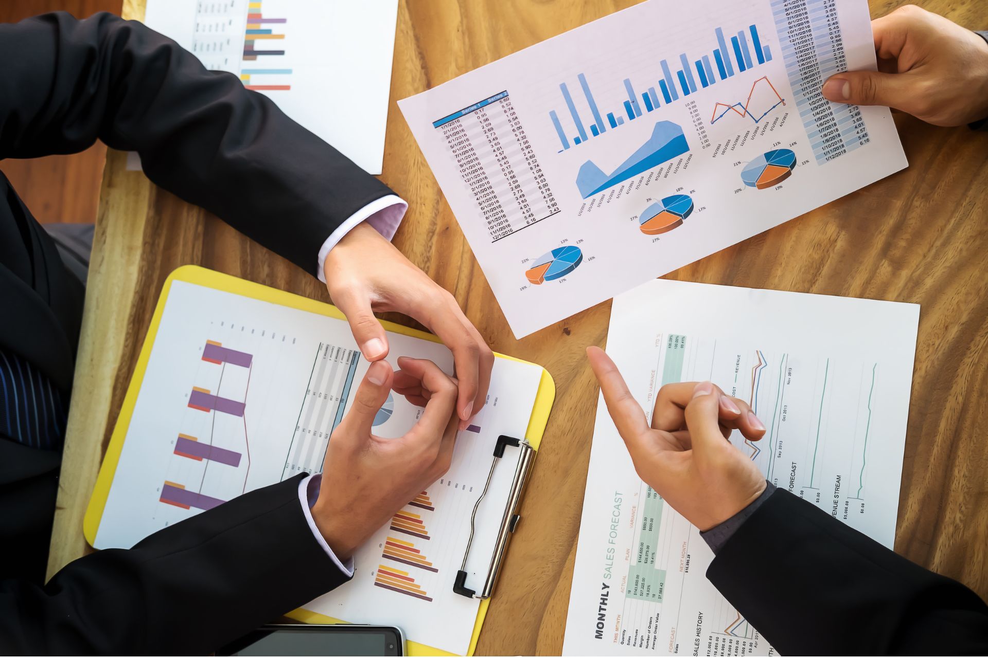 Two professionals reviewing financial charts and trade data reports on a wooden table during a business meeting.
